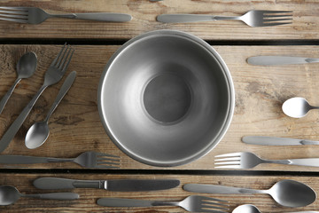 Metallic bowl and silver cutlery on wooden table, top view