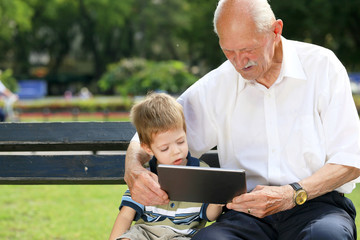 Grandchild teaching to his grandfather to use tablet on a bench