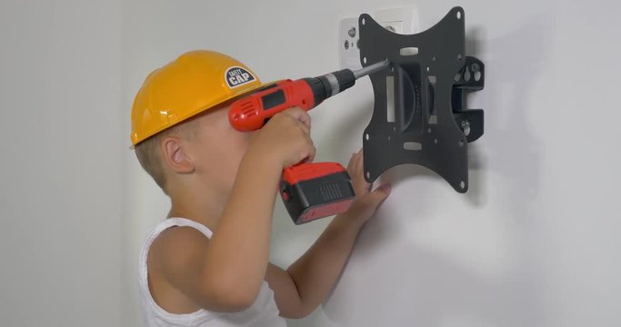 Little boy in safety helmet is fastening wall mount with electric screwdriver.