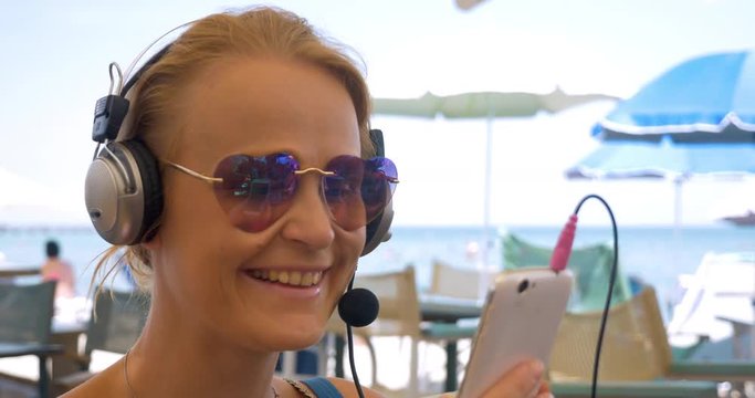 Close-up shot of young woman in heart-shaped sunglasses wearing headset and having a video chat on smart phone. Happy girl showing beach to the talker and blowing kiss finishing the chat