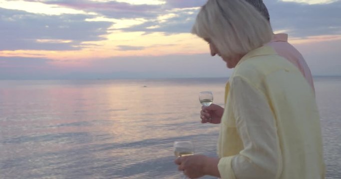 Steadicam shot of mature couple walking by the sea at sunset with wine glasses in hands.