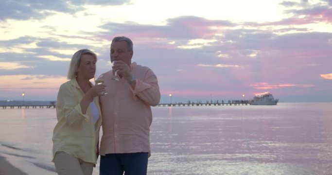 Steadicam shot of loving mature couple enjoying romantic evening on the seashore. They walking along the coast and having wine 