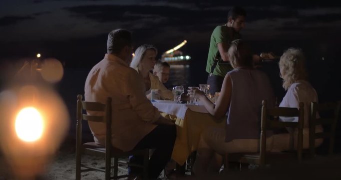 Family having meal in outdoor restaurant on the beach at night. Waiter serving dinner, burning tiki torch in foreground