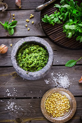 Pesto in marble mortar, salt, pine nuta and garlic on wooden table.