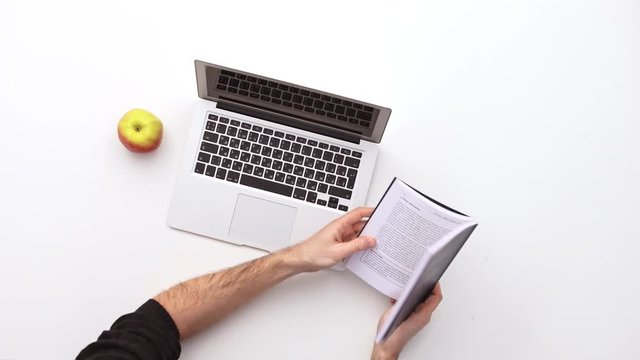 Man searching something in book surrounded by a laptop on his desk and apple.  Upview