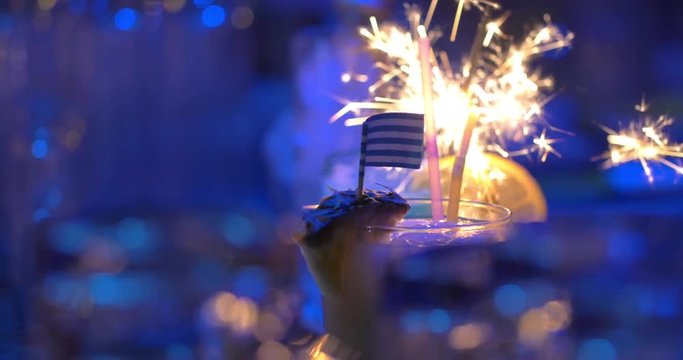 Close-up shot of two cocktails in the bar. Sparkler going down in one of them, another with pineapple and Greek flag