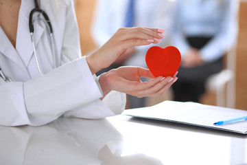 Female doctor with stethoscope holding heart.  Patients couple sitting in the background