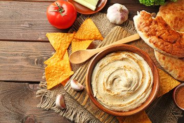 Wooden bowl of tasty hummus with chips and flat bread on table