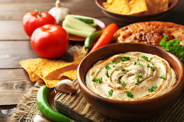 Wooden bowl of tasty hummus with chips and parsley on table