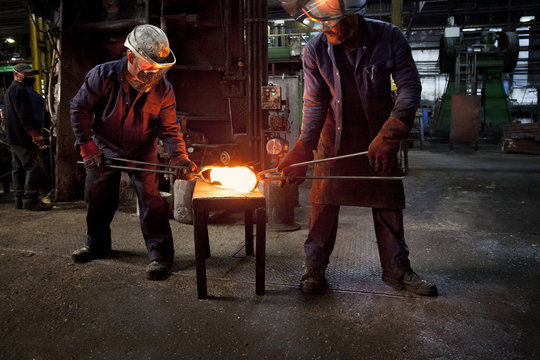Forge worker hands over pre formed steel billet to colleague for presentation to set of two dies in hammer to be fully forged into crank shaft