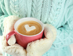 Female hands in warm mittens holding cup of hot cappuccino with heart marshmallow, close up