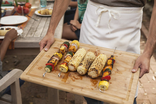 Man holding tray of barbecued vegetable skewers and corn on the cob