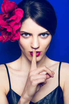 Attractive brunette young woman gesturing to be quiet. Isolated on a blue background with big red flower in hair.