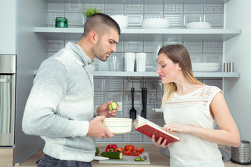 Obraz na płótnie Canvas Happy young couple is preparing healthy meal together in the kitchen. Young woman is reading recipe from the cookbook, and young man is trying to follow her instructions.