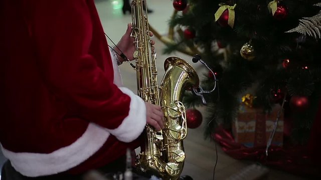 Saxophonist in Santa Claus suit playing merry Christmas carol near New Year tree