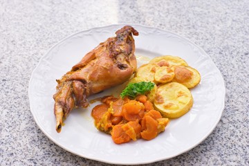 Gourmet roasted rabbit leg with glazed sweet carrot and baked potatoes choped on thin slices. Meal is served on the white simply plate and granit table isolated. Healty and light meal perfect for diet