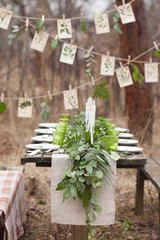 Weddind table setting with white plates and green glasses decorated with white candles, green leaves and eucalyptus and a garland of green leaves and postcards outdoors