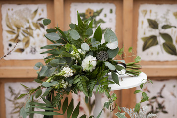 Green bouquet of eucalyptus, ivy, fern, aspidistra, ruskus, date fruit, pink, ranunkulyus with flax box, green and brown velvet ribbons on white stand and brown and green floral pattern on background