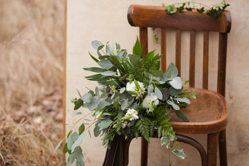 Green bouquet of eucalyptus, ivy, fern, aspidistra, ruskus, date fruit, pink, ranunkulyus with flax box, green and brown velvet ribbons on white stand and brown on wooden chair and beige background