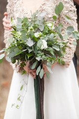Girl dressed in beige colours is holding green wedding bouquet of eucalyptus, ivy, fern, aspidistra, ruskus, date fruit, pink, ranunkulyus with green and brown velvet ribbons.