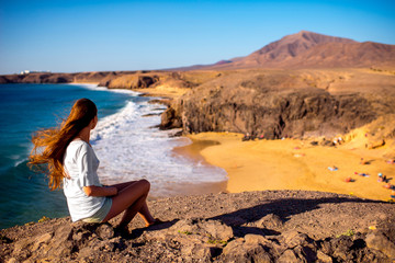 Young female tourist sitting and enjoying beautiful view on Papagayo beach on Lanzarote island in Spain - obrazy, fototapety, plakaty