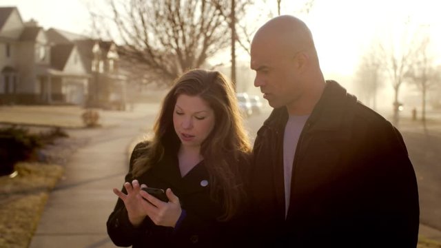 Couple using a smart phone while checking out a house (off screen) in a typical US Mid West suburban street.  Early morning sunlight with golden tint.  Cropped from 4K