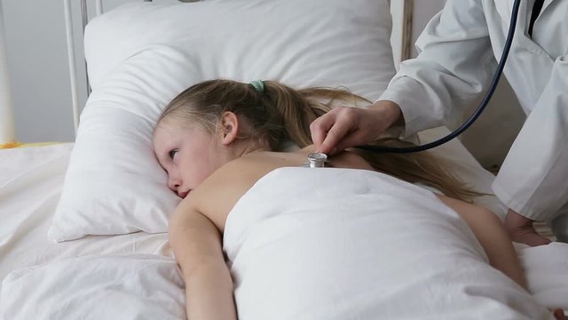 Woman doctor with a stethoscope auscultating to the child lying in the bed.Doctor or nurse examines the girl in the bed stethoscope.The doctor in a white room with a stethoscope.Woman doctor with a