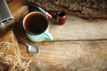 Metal mug of coffee with flask and tobacco pipe on wooden background