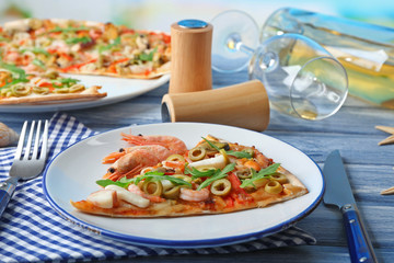 Pizza slice with seafood, red pepper, green olives and bottle of wine on blue wooden table