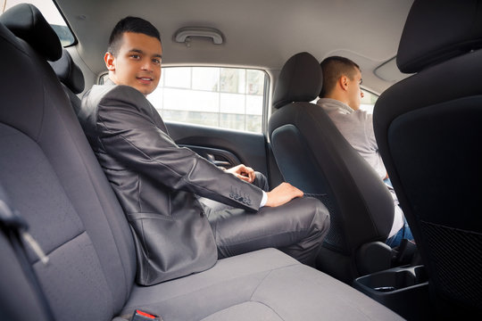 Young businessman is looking at camera while sitting on back seat of the car, while his chauffeur is driving automobile.