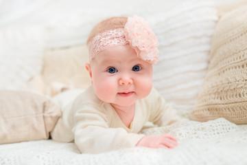 Smiling baby girl with chubby cheeks and big blue eyes wearing white clothes and pink band with flower lying on bed and looking at camera. Babyhood and childhood concept
