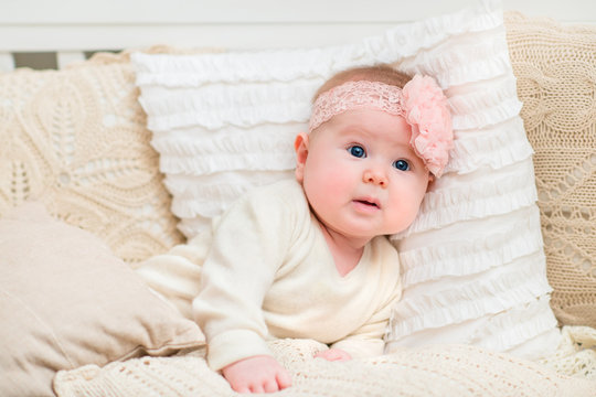 Surprised beautiful baby girl with chubby cheeks and big blue eyes wearing white clothes and pink band with flower lying on bed with knitted pillows. Babyhood and childhood concept