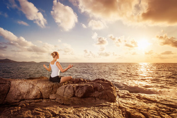 Young woman is training yoga meditating on a rock at sea sunrise