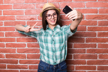 Beautiful young dark-haired girl in casual clothes and hat, eyeglasses, posing, smiling, making selfie, standing against brick wall