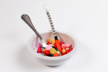 Many colored pills, capsules and syringe in a bowl with a spoon.