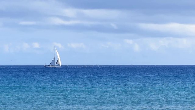 Grey white sailboat crossing the wide blue calm ocean. No waves and a partly cloudy sky.