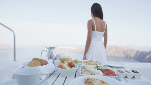 Food on Breakfast table with luxury travel woman looking at view. Variety of delicious food served on breakfast table on terrace. Lady on vacation holidays in Santorini, Greece.