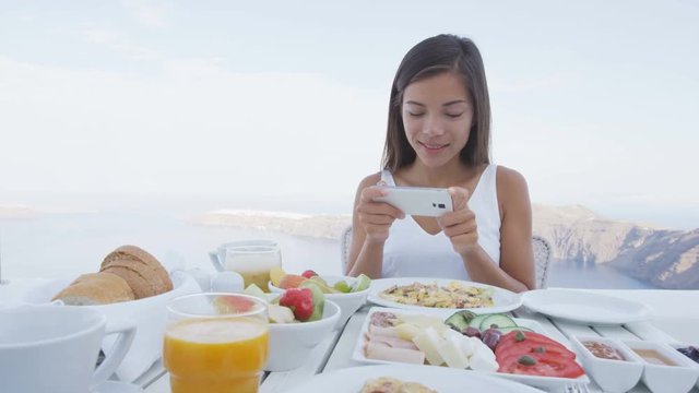 Beautiful woman using mobile phone app eating breakfast. Variety of healthy food items served on table. Female tourist is relaxing on terrace resort in Santorini.