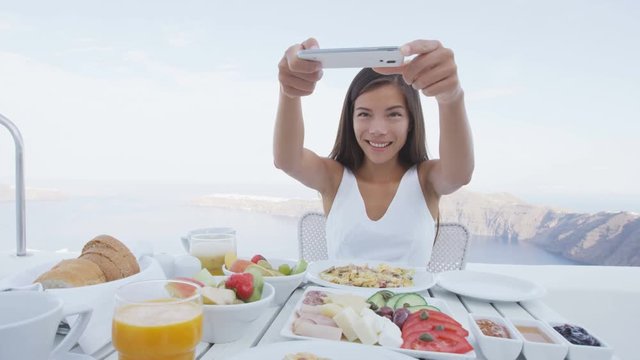 Woman taking pictures of breakfast using mobile cell smart phone app. Girl taking photo of food on luxury travel vacation for social media. Beautiful female in resort in Santorini, Greece, Europe.