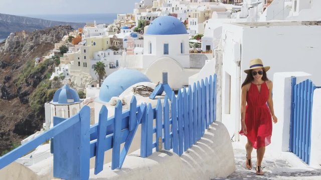 Travel tourist woman in Oia, Santorini, Greece. Happy young woman walking on stairs by famous blue dome church landmark destination. Beautiful girl in red dress on visiting the Greek island.