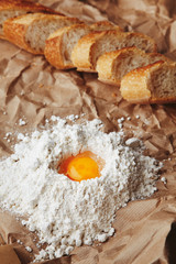 Obraz na płótnie Canvas Raw egg broken in white flour on craft paper near sliced french crunched freshly baked baguette bread. Yolk in center on flour pyramide