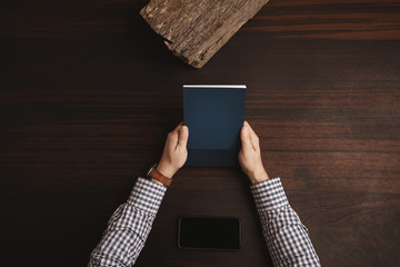 Accountant hands in plaid dress shirt holding closed note book above phablet lying horizontally in center. Isolated on red wooden table in meeting room 
