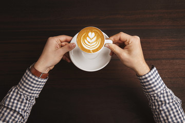 Businessman hands in plaid dress shirt wear luxury watches with leather strap hold white ceramic cup of cappuccino with foam coffee art above red wooden table in cafe shop