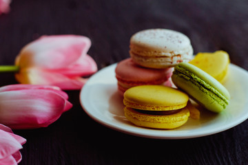 A beautiful flowers pink tulips with colorful macaroons laid on a white platter on dark wooden background