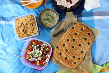 Top view of various picnic food: vegetable and feta salad, baba ghanoush, gluten-free crackers and olive bread.
