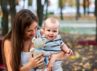 Happy mother and her baby-boy on hands play keeps teady bear toy outdoors in park.