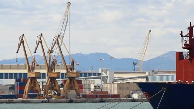 Port in front of mountains. Cranes and warehouses in the background, a container ship in the foreground. Transport crane carrying a white container.