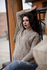 Spring in the country style. Full length portrait of young woman in white knitted sweater and furry hat standing in the front of rustic wood wall in the yard near house, wearing sweater casual.