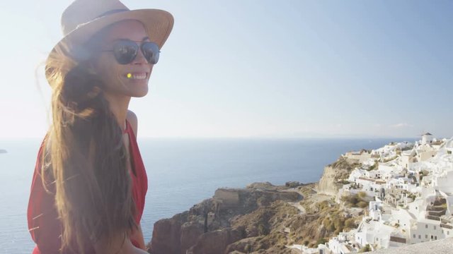 Smiling tourist woman enjoying beautiful view of village Oia and sea. Young female is wearing sunglasses and sunhat. She is enjoying her summer vacation in Santorini.