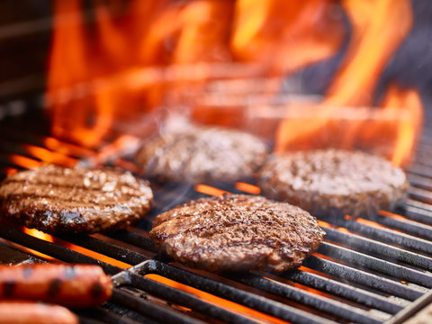hamburger patties cooking on flaming grill with hot dogs